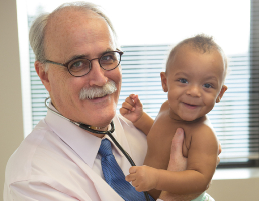 Dr. Murphy con estetoscopio, sonriendo y sosteniendo a un joven bebé sonriente con un tono de piel medio