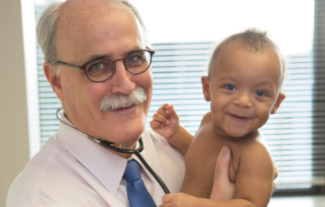 Dr. Murphy con estetoscopio, sonriendo y sosteniendo a un joven bebé sonriente con un tono de piel medio