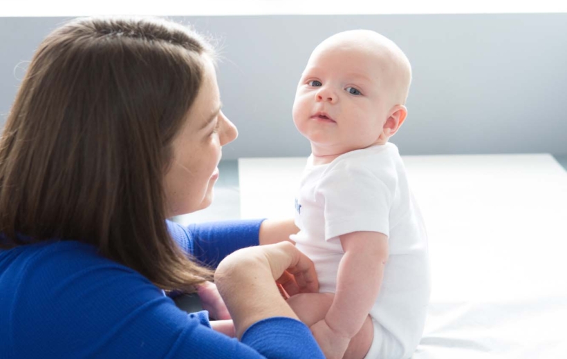 Mother with light skin, brown hair, wearing dark blue shirt, kneeling and interacting with baby sitting on exam table. Baby is dressed in white and looking at the camera.