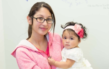 Woman with glasses, smiling slightly, wearing pink jacket and holding young girl wearing white dress and headband with flowers. Both have medium skin and dark hair and are looking into the camera.