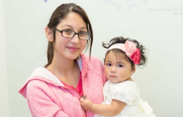 Mujer con gafas, ligeramente sonriente, lleva una chaqueta rosa y sostiene a una joven con vestido blanco y diadema con flores. Ambas tienen la piel mediana y el pelo oscuro y miran a la cámara.