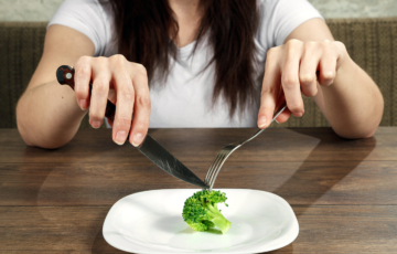 Photo of torso and hands of a person sitting at a table with a knife and fork cutting into a single piece of broccoli on a white plate.