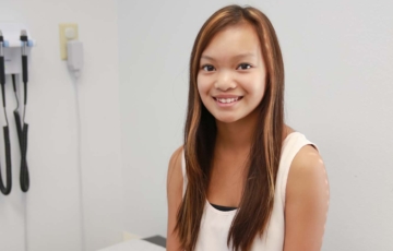 smiling teen girl sitting on exam table with long brown hair, white shirt, and jeans